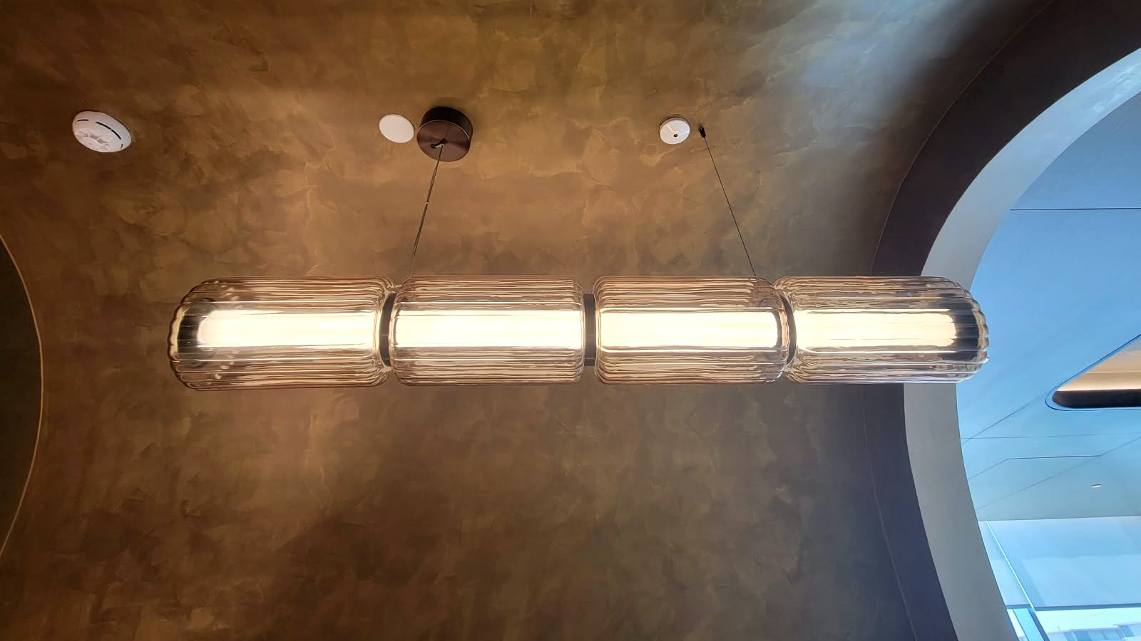 Four fluted glass modules joined into a lit linear run beneath a bronze plaster vaulted ceiling
