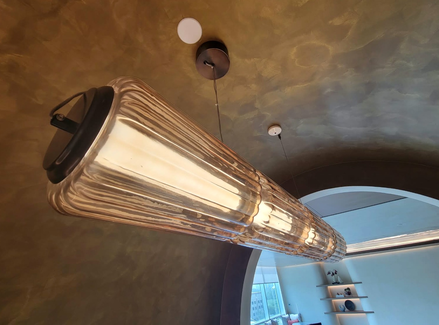 Fluted glass pendant seen end on, lit, beneath a bronze plaster vault